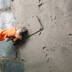 Mano con guante naranja aplica enfoscado rugoso a pared de bloques de cemento con llana.