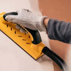 Mano enguantada usando una lijadora amarilla para lijar paredes, preparando la superficie para pintar.