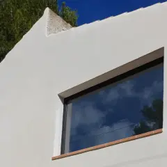 Fachada blanca con mortero de cal, reflejando el cielo azul y nubes. Ventana moderna y árbol verde.