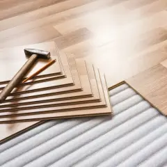 Tablas de suelo laminado apiladas con martillo y lápiz, listas para instalar sobre una base que protege de la humedad que sube del suelo.