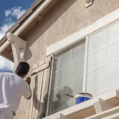 Hombre pintando persianas de madera para el acabado fachada de una casa. Cubo de pintura azul y blanca a la vista.