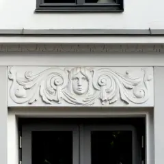 Detalle arquitectónico: una cornisa con un rostro femenino y volutas decorativas sobre una puerta.