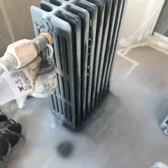 Proceso de pintar radiadores de hierro con pistola. Guante protege mano mientras se aplica pintura gris.