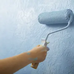 Mano pintando una pared con rodillo azul. El plástico del rodillo se ve salpicado de pintura.