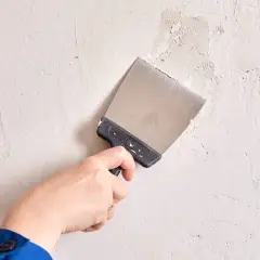 Mano con espátula metálica trabajando en la pared para alisar paredes y reparar imperfecciones.