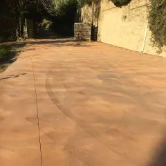 Un camino de **suelo hormigon pintado** de color terracota, con marcas de neumáticos, conduce a una puerta abierta.