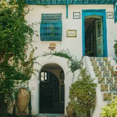 Fachadas antiguas con escaleras de azulejos coloridos, puerta azul y vegetación exuberante. Un portal arqueado y vasijas de barro completan la escena.