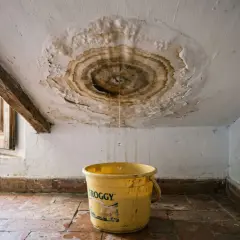 Goteras en el techo, una cubeta amarilla recoge el agua. Urge reparar tejado sin quitar tejas.