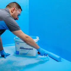 Hombre aplica látex azul con rodillo, creando sistemas de impermeabilización en paredes y suelo.