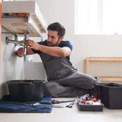 Fontanero trabajando en el lavabo, listo para cambiar tuberías en casa. Herramientas y cubos a su lado.