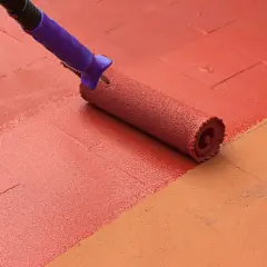 Rodillo aplicando pintura para suelo de cemento de color rojo sobre una superficie con baldosas de color naranja.