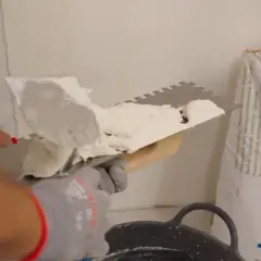 Man applying morteros para fachadas con llana dentada. Bolsa de producto de construcción al fondo.