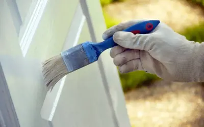 Mano enguantada aplicando pintura blanca a una puerta con brocha. Proceso de lacar puertas en blanco para renovar su aspecto.