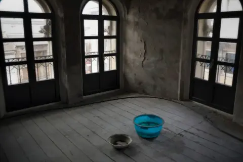 Habitación con tres ventanas arqueadas y suelo de madera. Hay dos cubos en el suelo, uno azul y otro gris, recogiendo agua. Se puede caer el techo por una gotera.