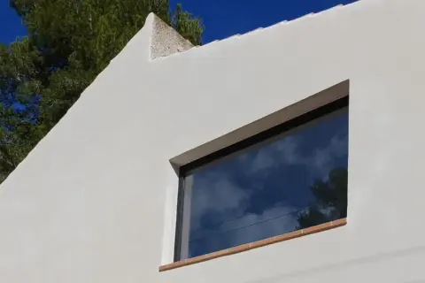 Fachada blanca con mortero de cal, reflejando el cielo azul y nubes. Ventana moderna y árbol verde.