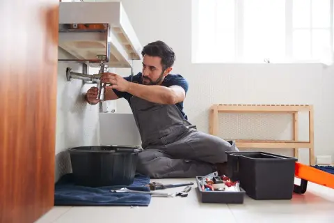 Fontanero trabajando en el lavabo, listo para cambiar tuberías en casa. Herramientas y cubos a su lado.