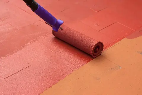 Rodillo aplicando pintura para suelo de cemento de color rojo sobre una superficie con baldosas de color naranja.