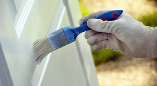 Mano enguantada aplicando pintura blanca a una puerta con brocha. Proceso de lacar puertas en blanco para renovar su aspecto.