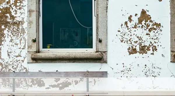 Paredes con desconchones y manchas de humedad, evidencia de filtraciones de agua de lluvia. Una ventana refleja el interior y un banco se sitúa delante.