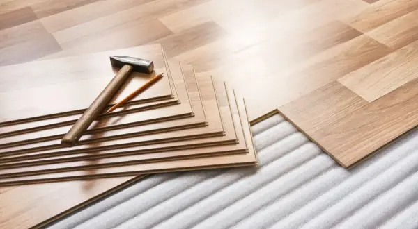 Tablas de suelo laminado apiladas con martillo y lápiz, listas para instalar sobre una base que protege de la humedad que sube del suelo.