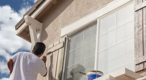 Hombre pintando persianas de madera para el acabado fachada de una casa. Cubo de pintura azul y blanca a la vista.