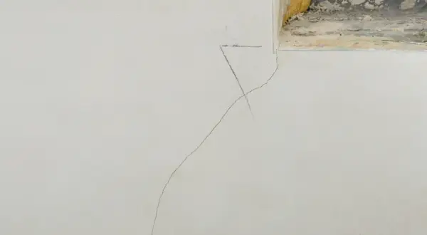 Grieta fina en pared de yeso, indicando la necesidad de reparar grietas por asentamiento. Se observa una marca de lápiz cerca de la grieta.