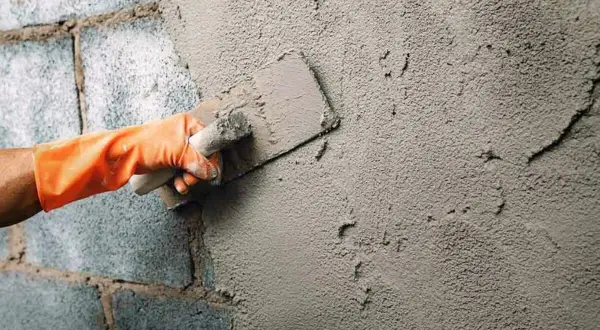 Mano con guante naranja aplica cemento a una fachada enfoscada con llana.
