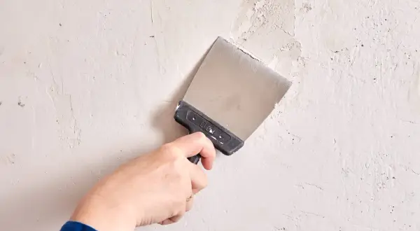 Mano con espátula metálica trabajando en la pared para alisar paredes y reparar imperfecciones.