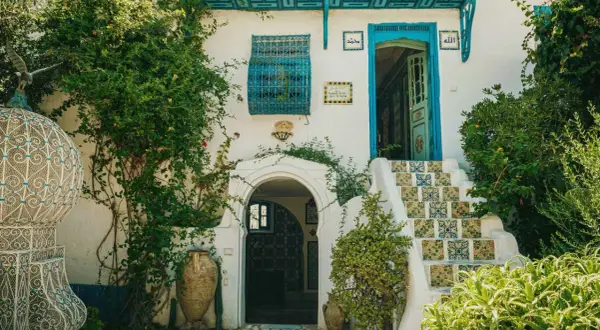 Fachadas antiguas con escaleras de azulejos coloridos, puerta azul y vegetación exuberante. Un portal arqueado y vasijas de barro completan la escena.