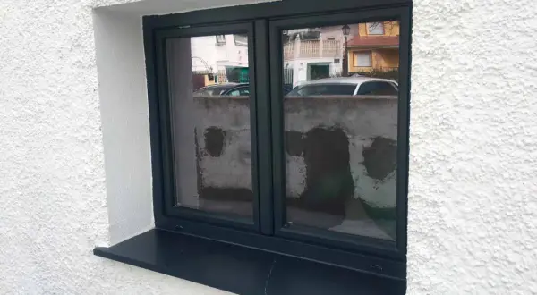 Ventana con marco oscuro y alféizar negro, lista para rematar ventanas por fuera. Refleja una calle con casas y coches.
