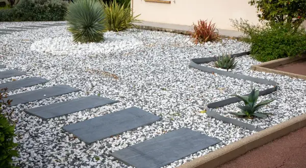 Senderos de piedra gris serpentean entre jardines con grava y piedras blancas y oscuras. Plantas suculentas y un gran arbusto verde completan el diseño.