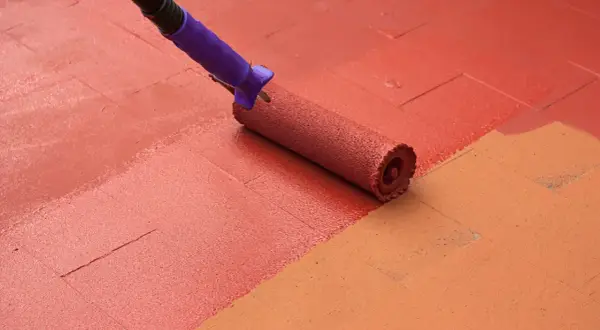 Rodillo aplicando pintura para suelo de cemento de color rojo sobre una superficie con baldosas de color naranja.