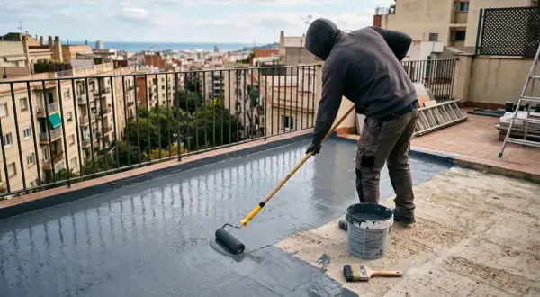 Hombre aplicando impermeabilizante a rodillo en terraza de 100 metros, con vistas a la ciudad y al mar.
