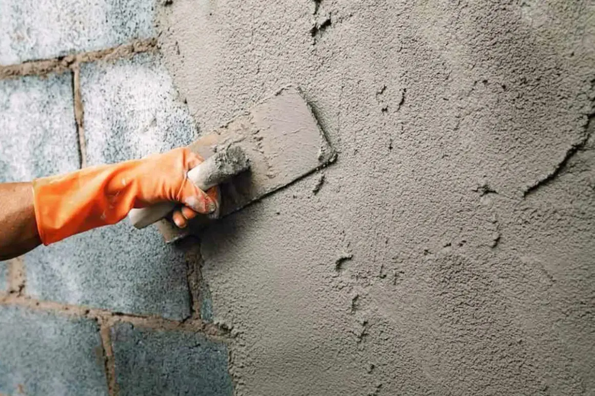 Mano con guante naranja aplica enfoscado rugoso a pared de bloques de cemento con llana.