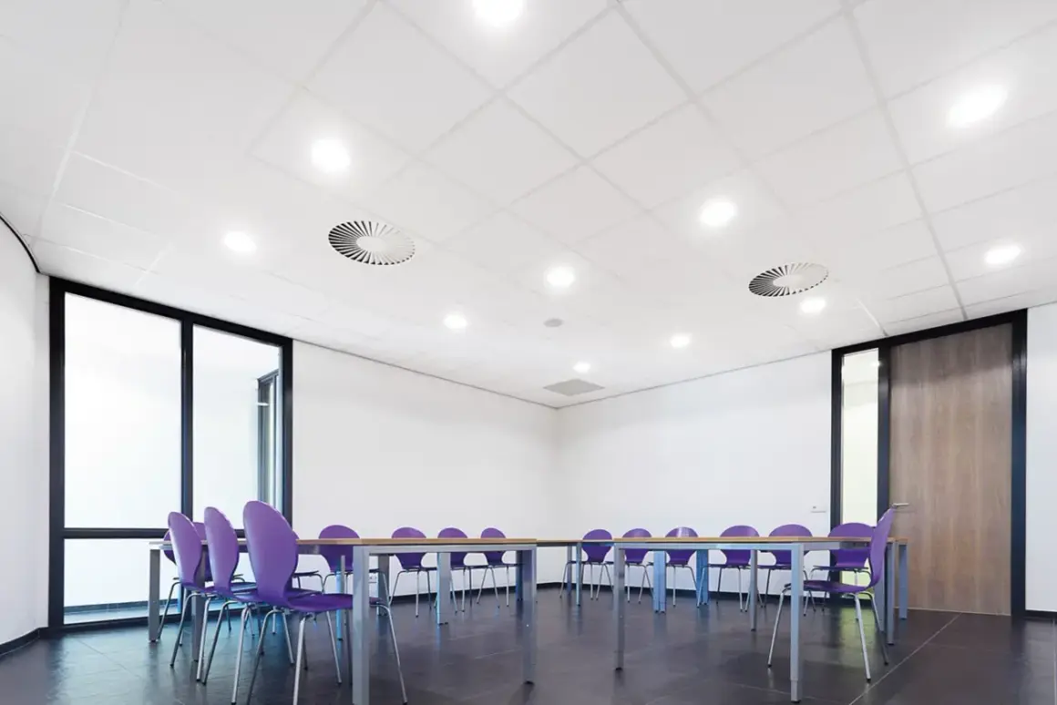Sala de reuniones con sillas moradas y mesa. El falso techo blanco con luces empotradas y rejillas de ventilación crea un ambiente moderno.