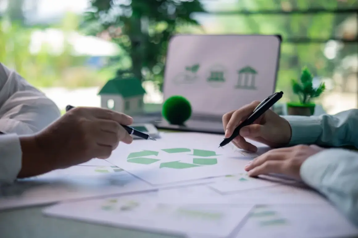 Manos revisando documentos con símbolo de reciclaje y un portátil con iconos de sostenibilidad, preparándose para una certificación verde.