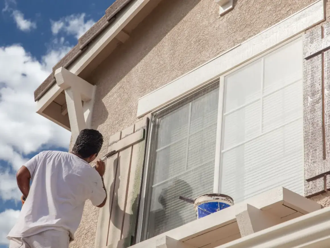 Hombre pintando persianas de madera para el acabado fachada de una casa. Cubo de pintura azul y blanca a la vista.