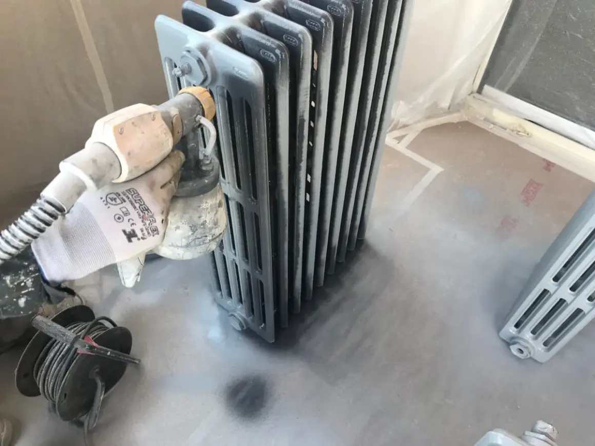 Proceso de pintar radiadores de hierro con pistola. Guante protege mano mientras se aplica pintura gris.