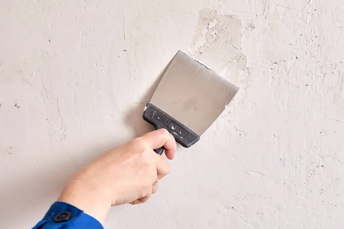 Mano con espátula metálica trabajando en la pared para alisar paredes y reparar imperfecciones.