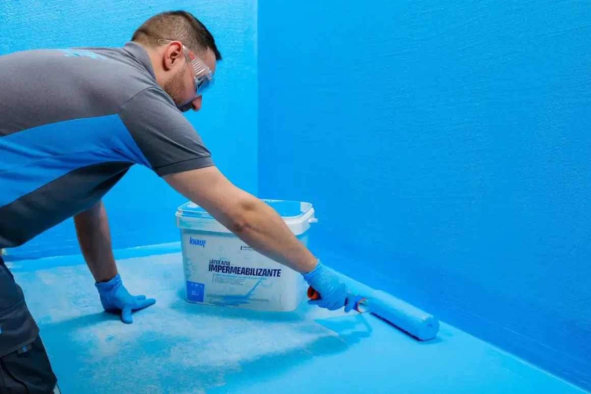 Hombre aplica látex azul con rodillo, creando sistemas de impermeabilización en paredes y suelo.