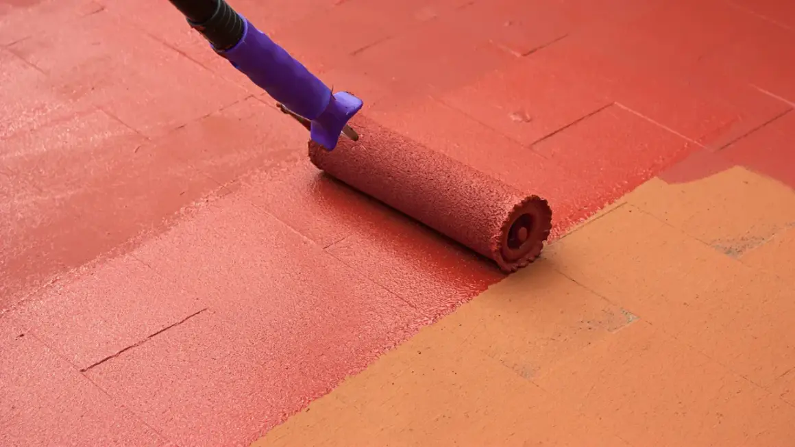 Rodillo aplicando pintura para suelo de cemento de color rojo sobre una superficie con baldosas de color naranja.