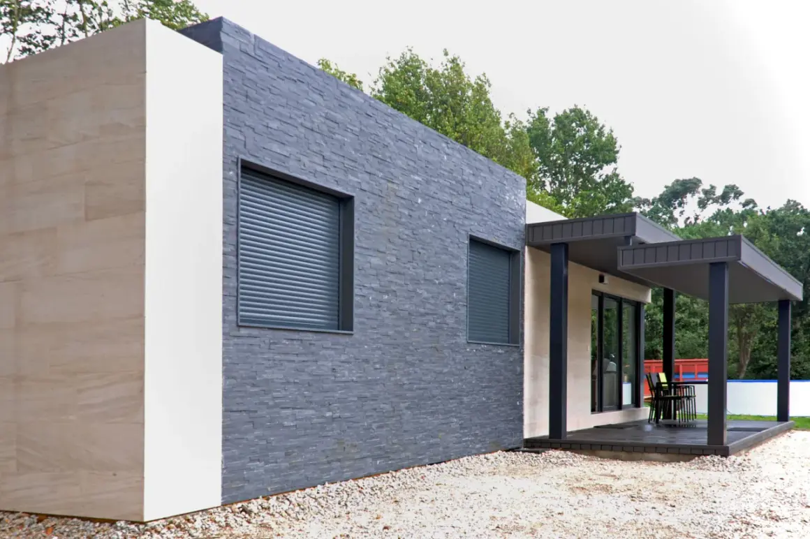 Muros exteriores de casas modernas con texturas de piedra y acabados claros. Ventanas con persianas enrollables y un porche con mobiliario.