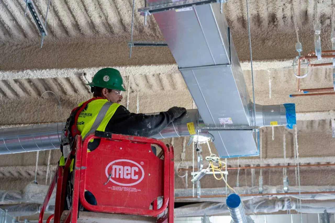 Trabajador en plataforma elevadora instala conductos de ventilación, como si fueran las venas de edificios enfermos que necesitan ser reparados.
