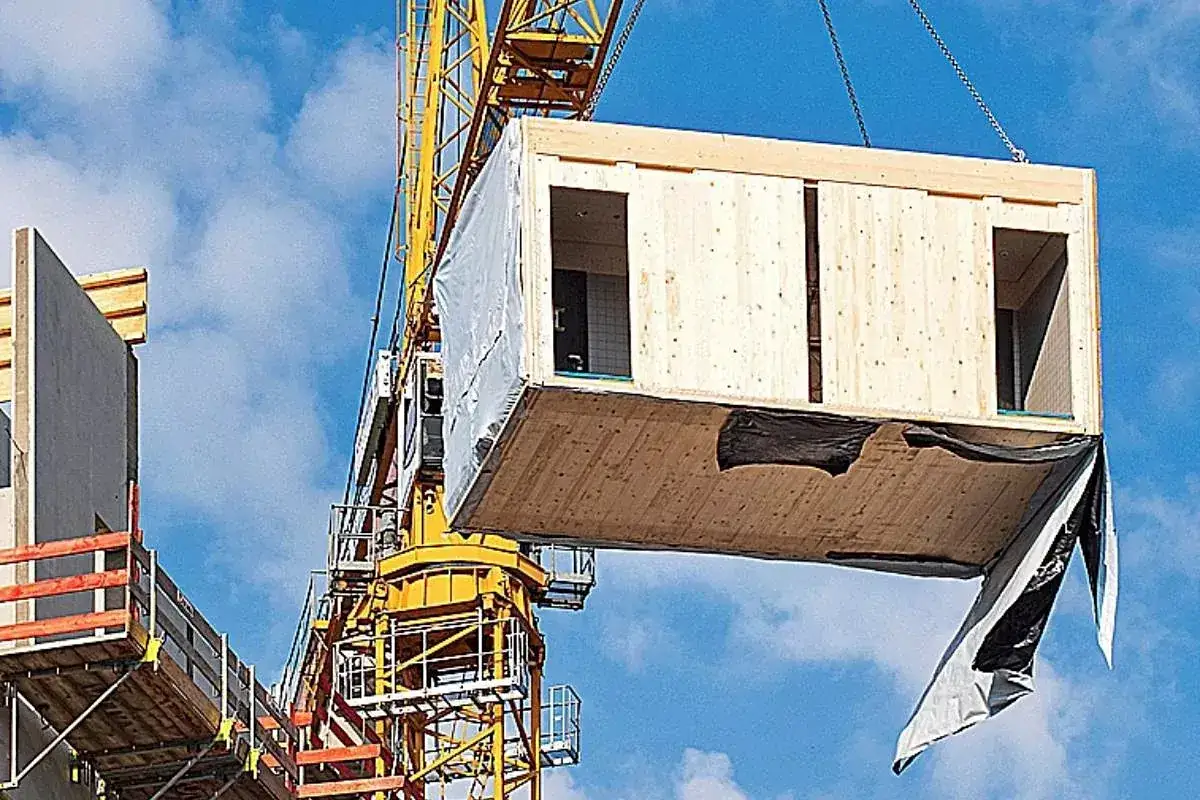 Grúa amarilla izando un módulo de construcción modular de madera. El módulo tiene aberturas para puertas y ventanas.