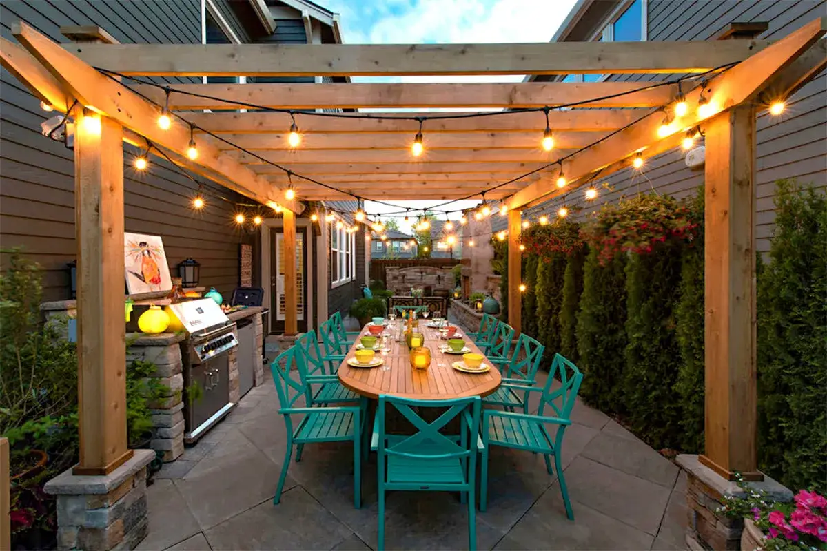 Ideas para iluminar una pérgola: luces de cadena crean un ambiente cálido sobre una mesa de comedor exterior y parrilla.