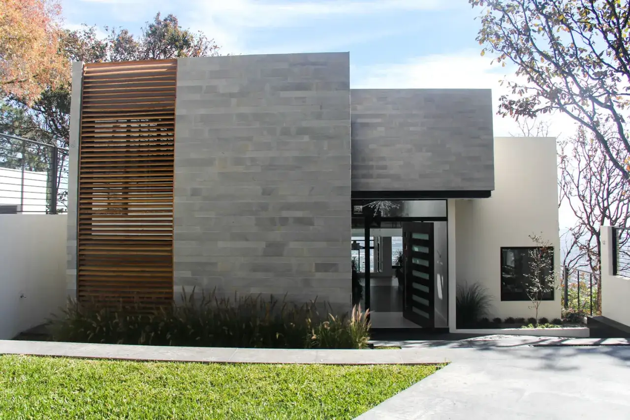 Modernos sistemas de fachadas combinan piedra gris y madera en esta casa, con una entrada abierta y vegetación.