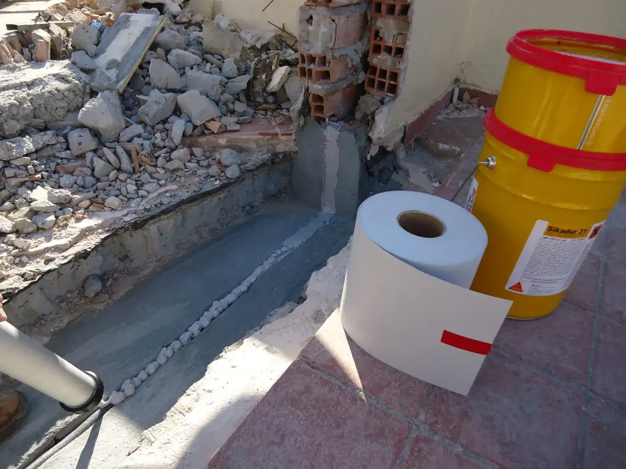 Aplicando sellador para juntas en una zanja de construcción. Cubos amarillos y rollo de material de impermeabilización cerca.