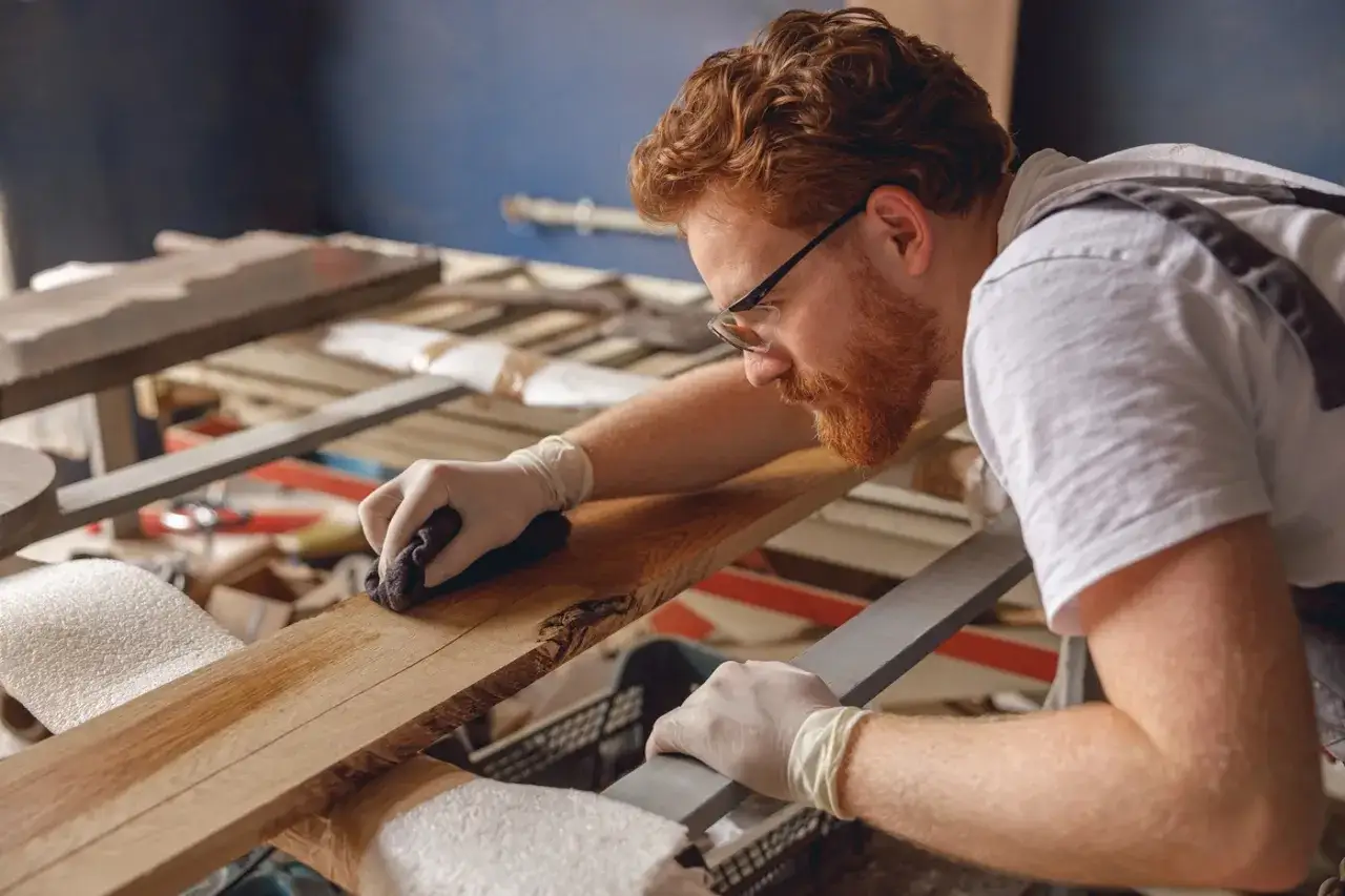 Artesano con barba pelirroja aplica producto para lacado muebles en madera.