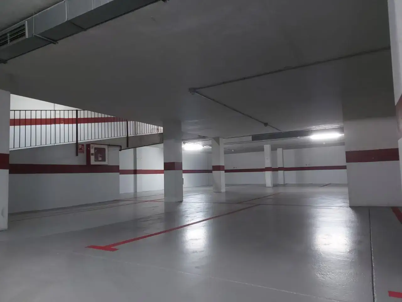 Amplio aparcamiento subterráneo con suelo de hormigón pintado de gris y líneas rojas. Pilares blancos y barandilla metálica.