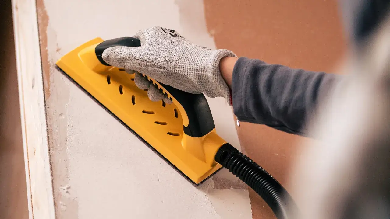 Mano enguantada usa una lijadora amarilla para alisar una pared.